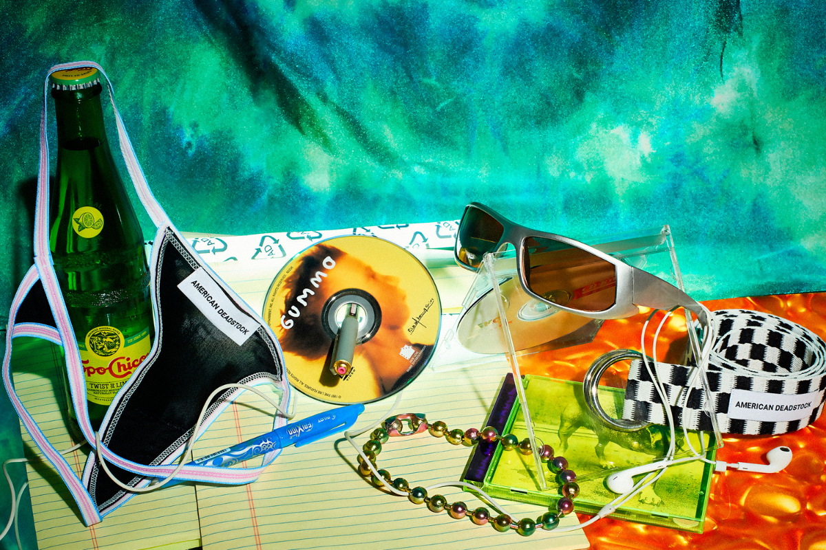 American deadstock tabletop image shot by Steve Harwick featuring a whale tail thong panty, oil slick ballchain choker, check it belt and silver y2k shield sunglasses. Shot on a blue green tie dye background with neon light, gummo cd, pens, paper and soda