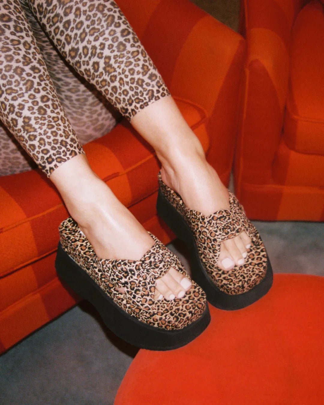 Leopard print platform slide sandals with thick black soles worn with matching leggings on a red chair.