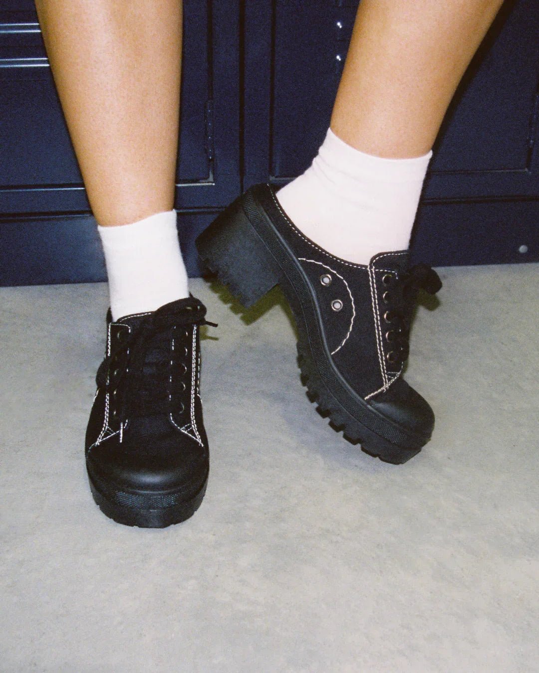 Black platform lace-up sneakers with white stitching worn with white socks on a light floor.