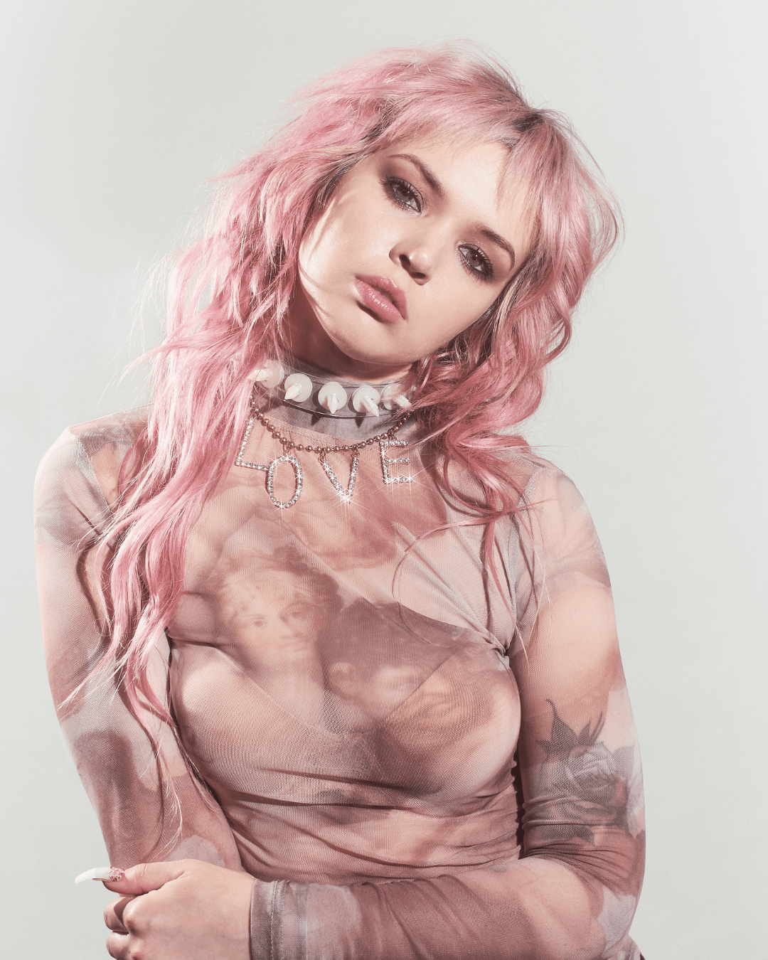 Woman with long pink hair wearing a sheer printed top and a silver LOVE letter necklace