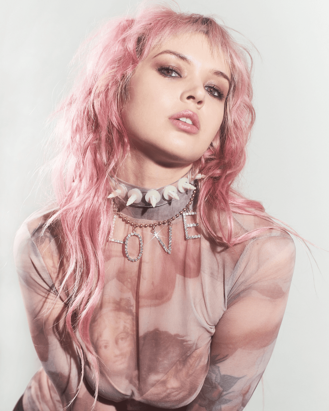 Woman with pink hair wearing a sheer printed top and a rhinestone 'LOVE' necklace with a white choker.