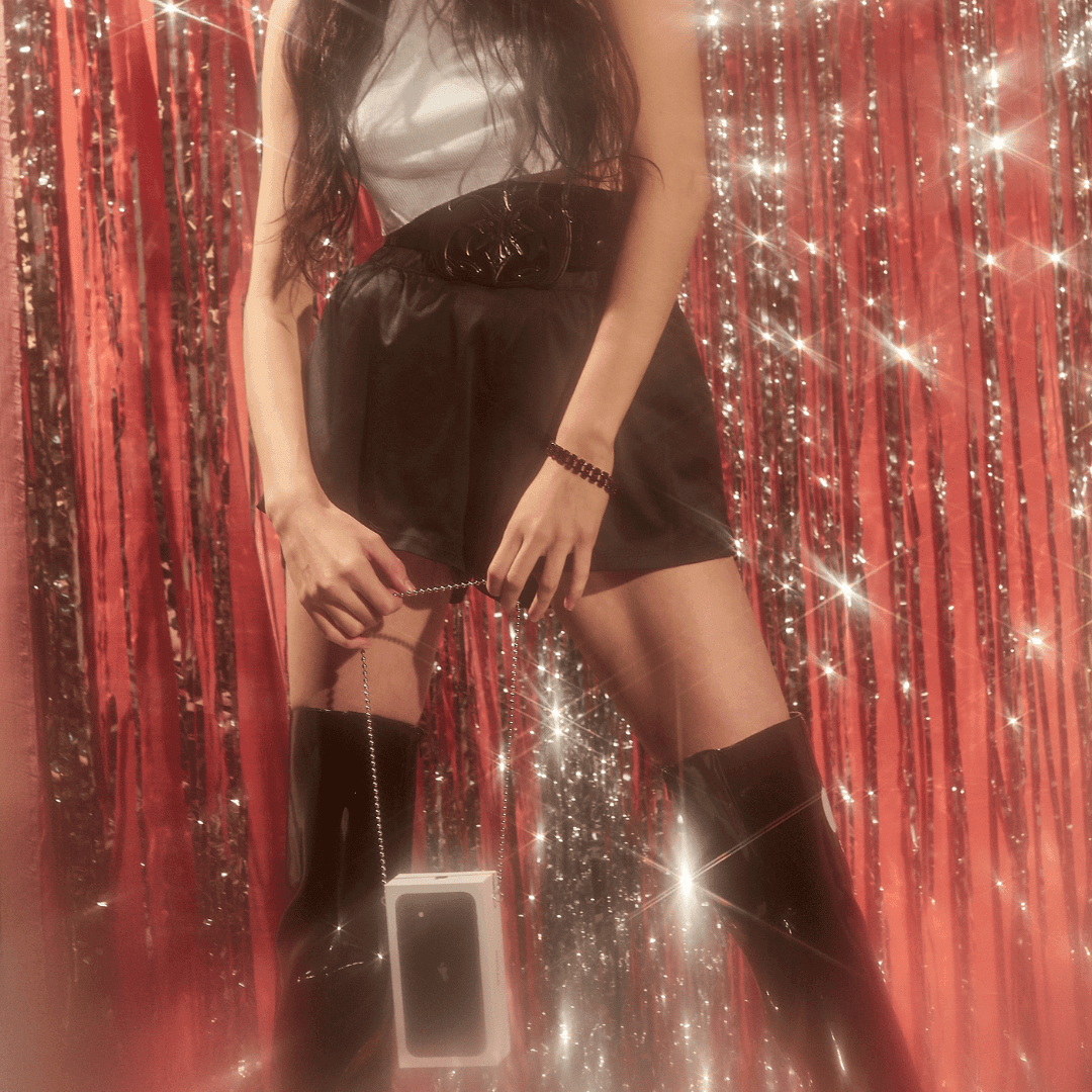 Woman wearing a white top, black skirt, black wide belt, and black boots with a phone case purse on a chain, standing against a sparkly red tinsel curtain background.