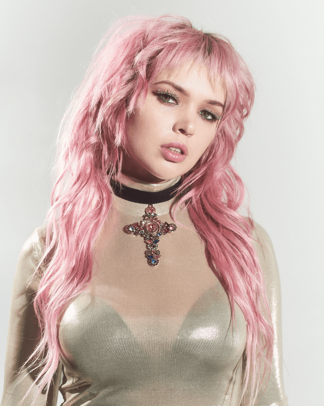 Woman with long pink hair wearing a sheer beige top and a black velvet choker with jeweled cross pendant