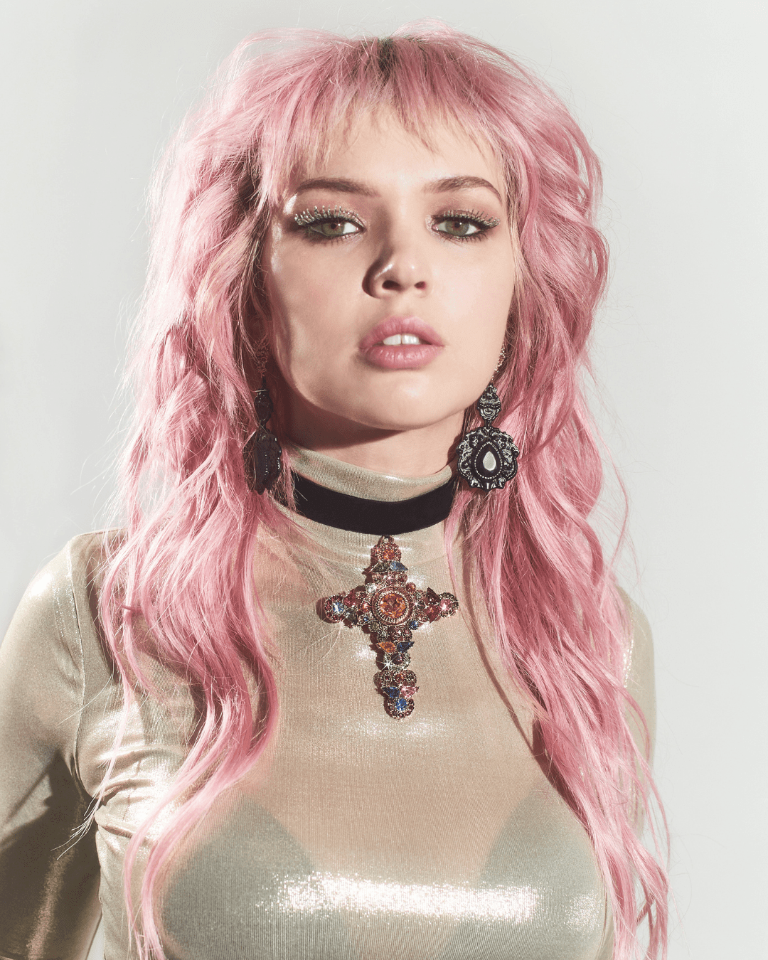 Woman wearing a black velvet choker with a large jeweled cross pendant, metallic top, and drop earrings