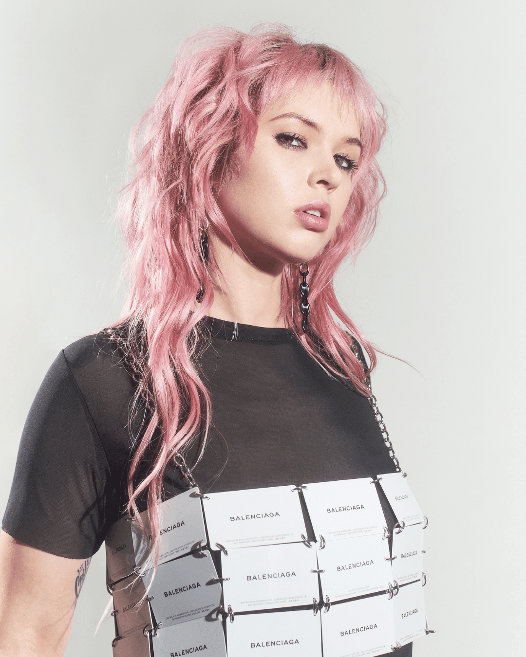 Person with wavy pink hair wearing a sheer black top and rectangular card-covered vest
