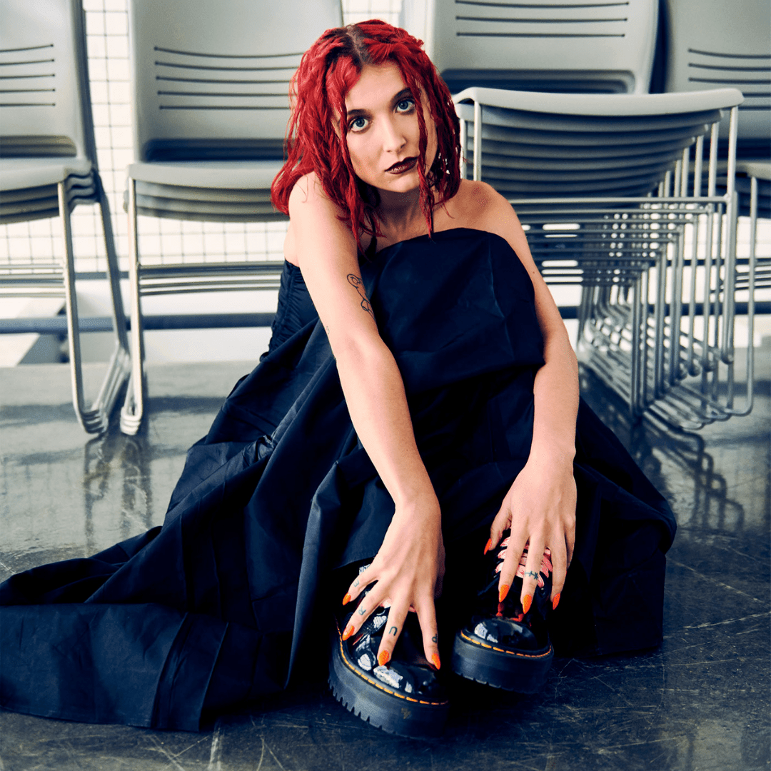 Person with red hair wearing a long black skirt and black platform shoes sitting on the floor
