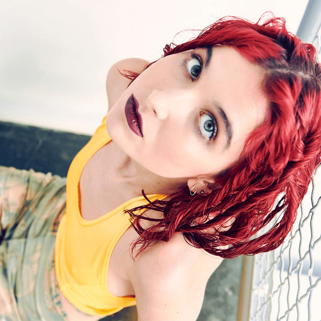 Person with red hair wearing a bright orange halter top, standing near a fence