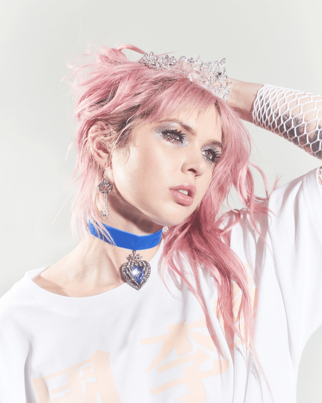 Woman with pink hair wears silver tiara, blue velvet choker with heart pendant, key earring, and white mesh sleeve.