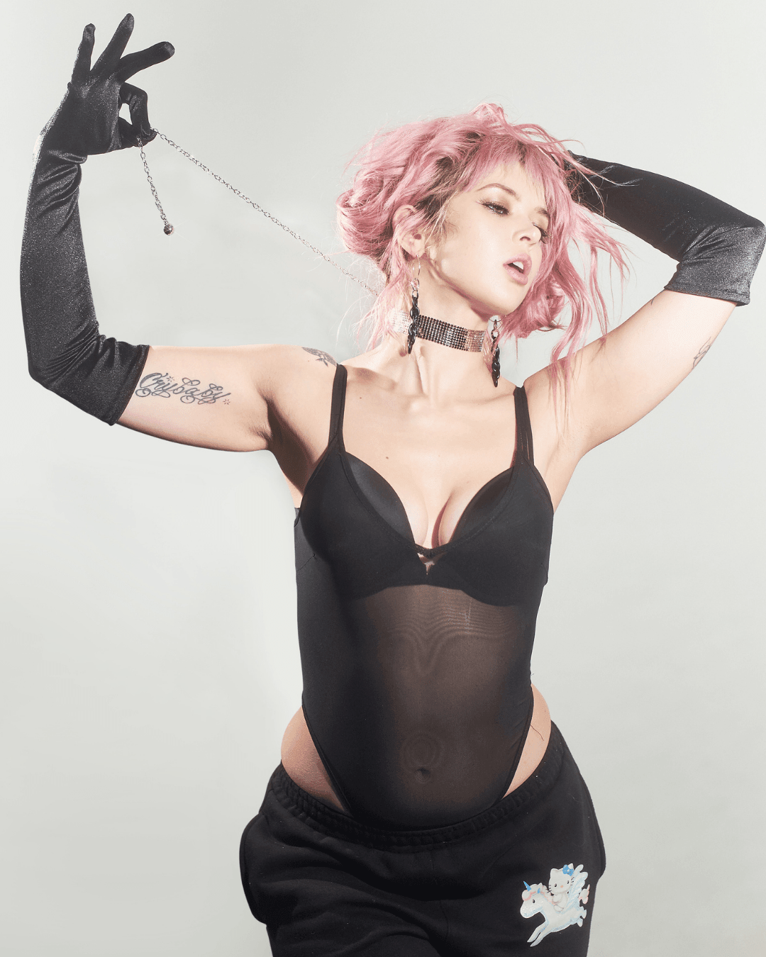 Woman wearing black mesh bodysuit, black chainmail choker, long black gloves, and black pants with a cartoon patch, standing against a light background.