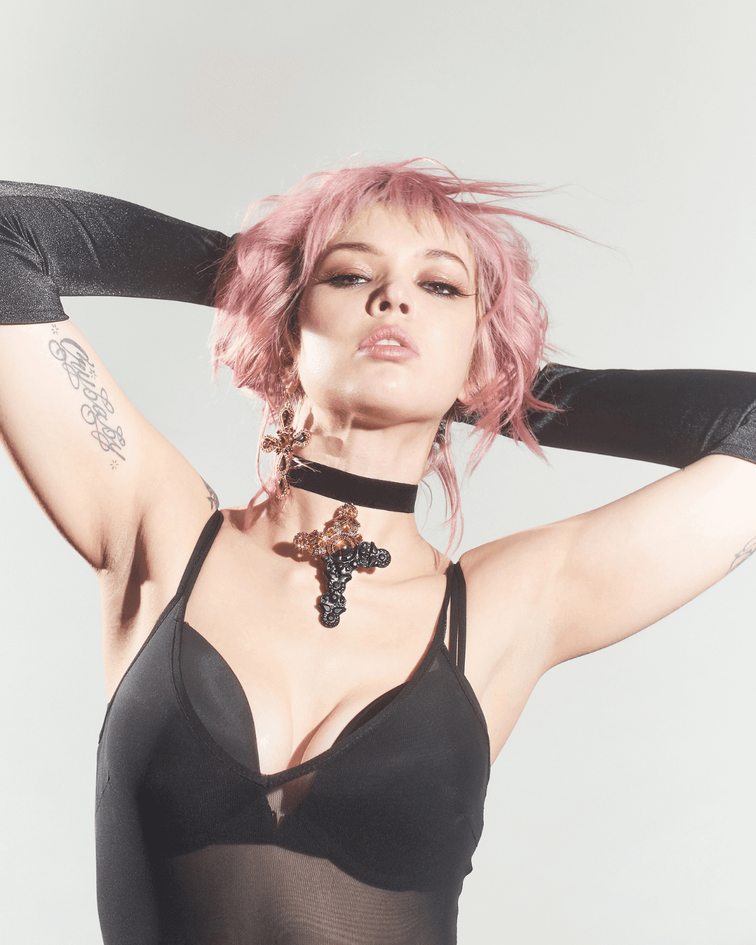 Woman with pink hair wearing a black mesh top, black velvet choker with large cross pendant, and long black gloves