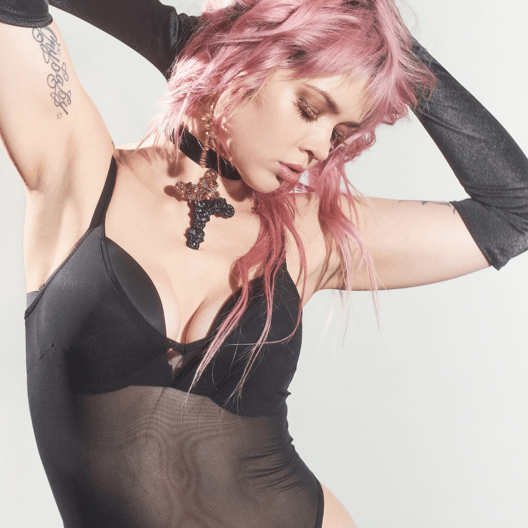 Woman wearing a black mesh bodysuit and long black gloves, with pink hair and a cross necklace