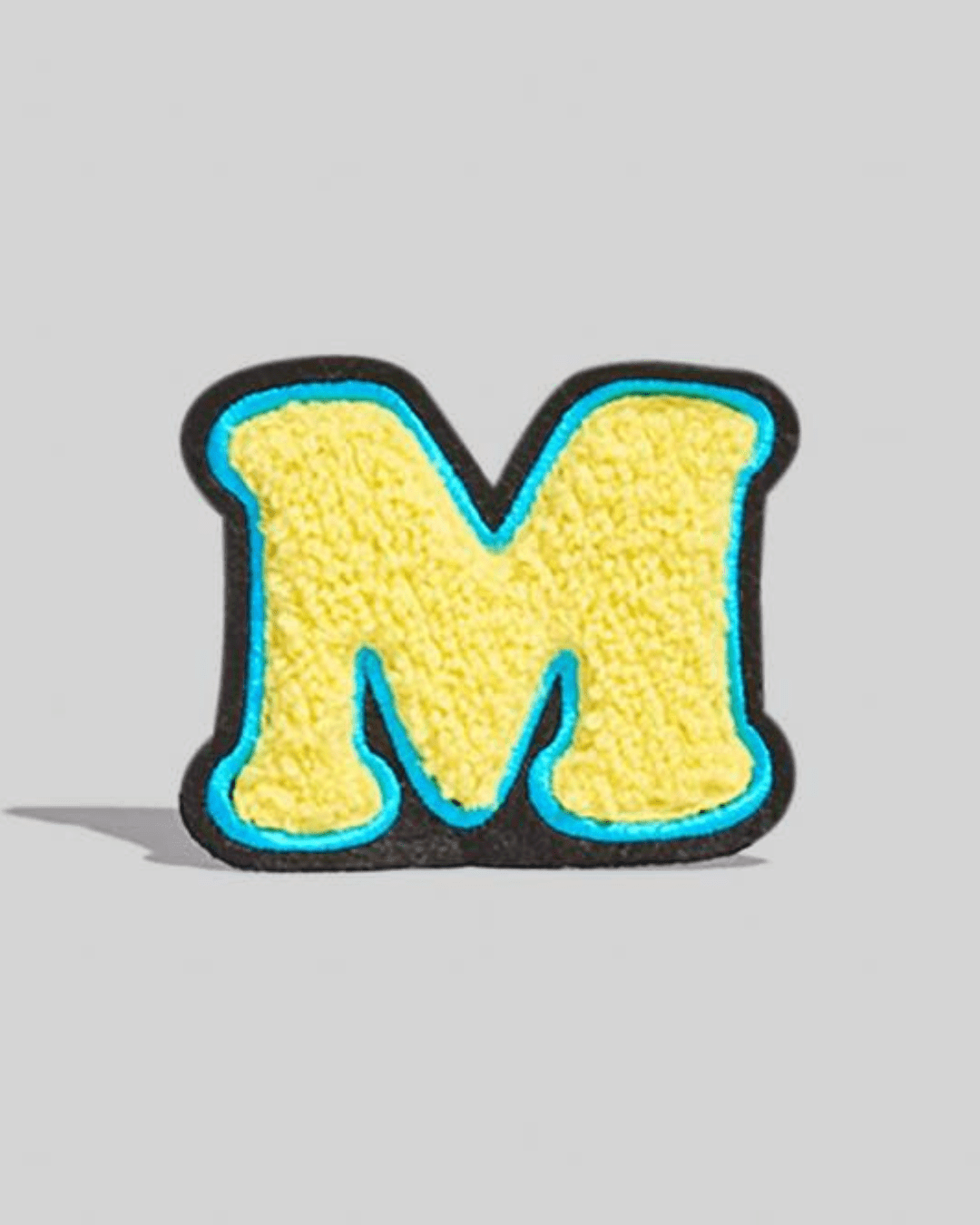 yellow chenille varsity letter M patch with blue and black border on a gray background